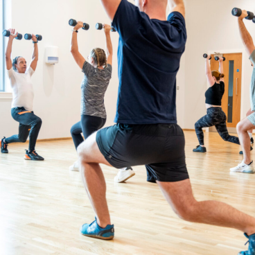 Group of people performing single-leg squats while holding small dumbbells overhead in a fitness class